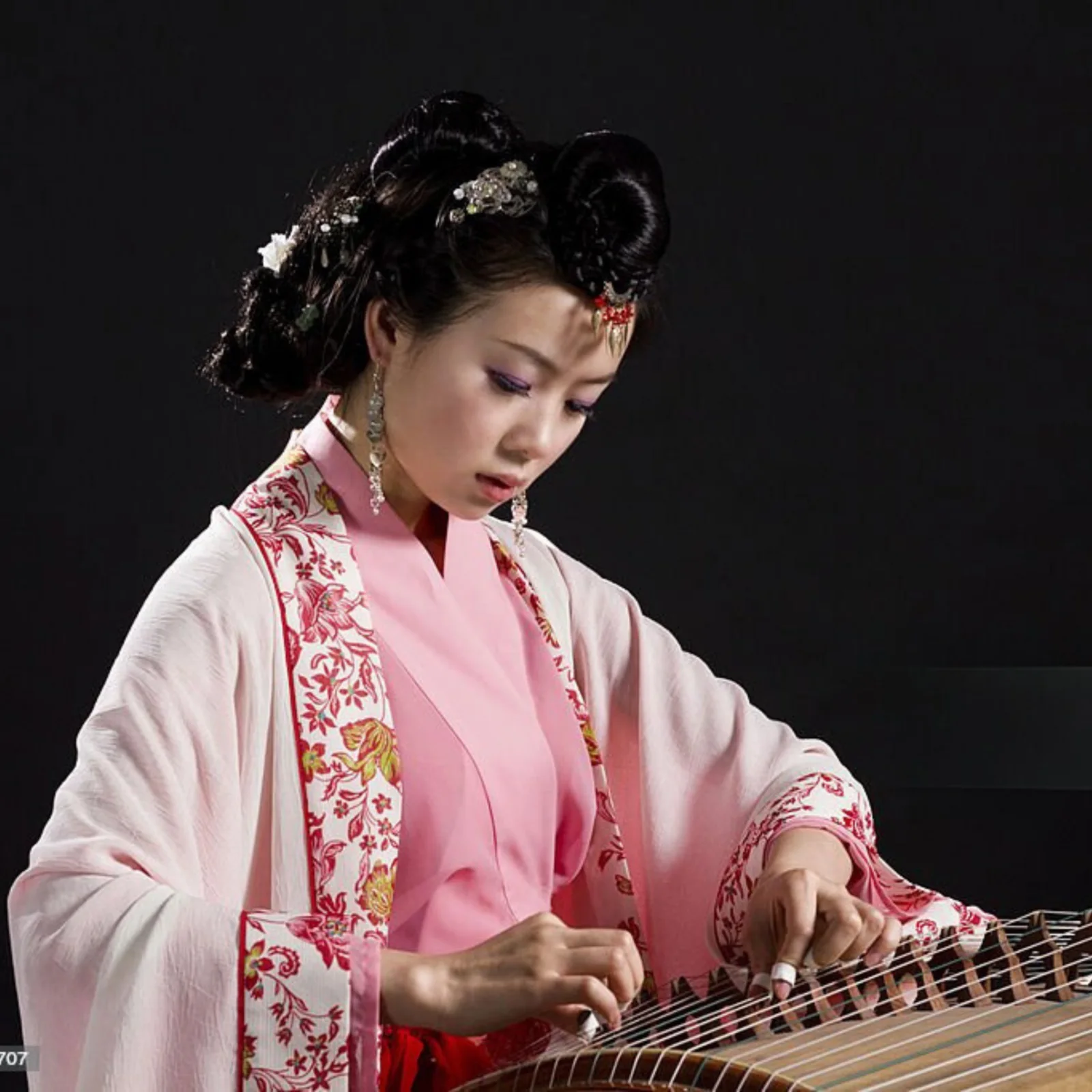 Người chơi đàn mặc cổ phục hồng viền hoa đỏ, cài trâm, gảy cây cổ tranh trên nền tối
