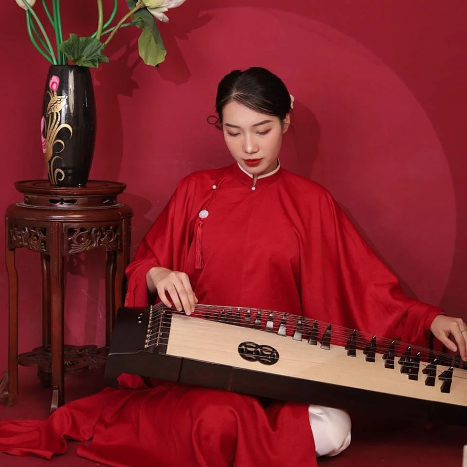 Người chơi đàn mặc áo dài đỏ ngồi gảy đàn tranh Việt Nam, bên bình hoa sen trên kỷ gỗ