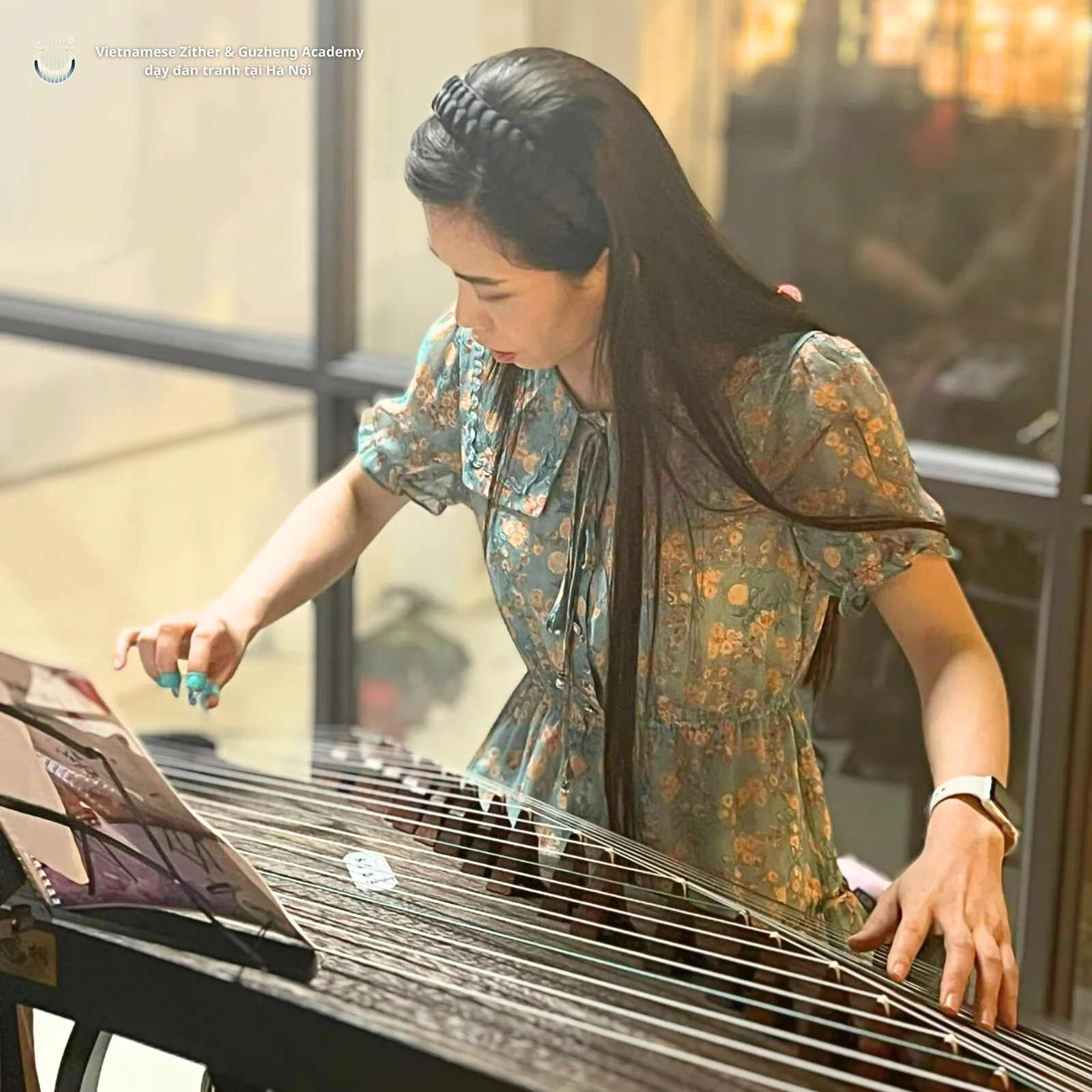 Người chơi đàn mặc váy hoa xanh, đeo móng gảy, chơi cổ tranh bên ô cửa kính