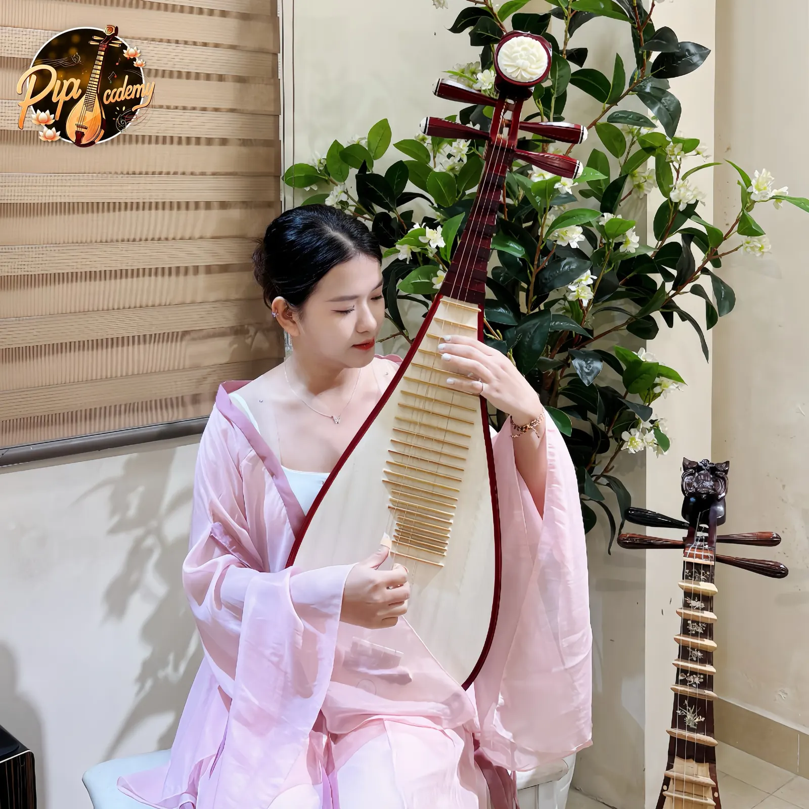 Người chơi đàn mặc áo lụa hồng nhắm mắt gảy cây đàn tỳ bà viền đỏ, bên chậu cây hoa trắng