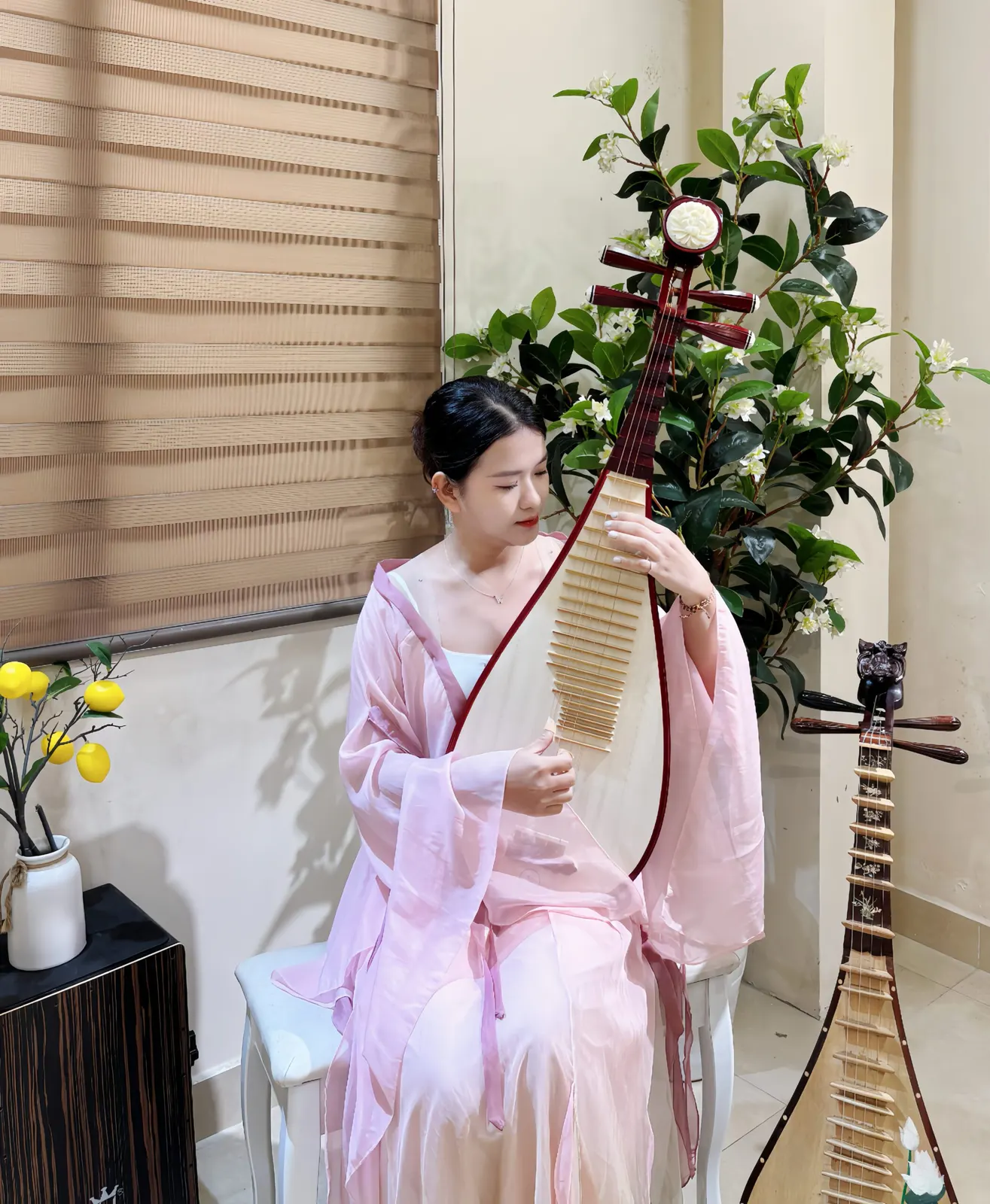 Người chơi đàn mặc áo lụa hồng, nhắm mắt gảy cây đàn tỳ bà mặt gỗ sáng viền đỏ