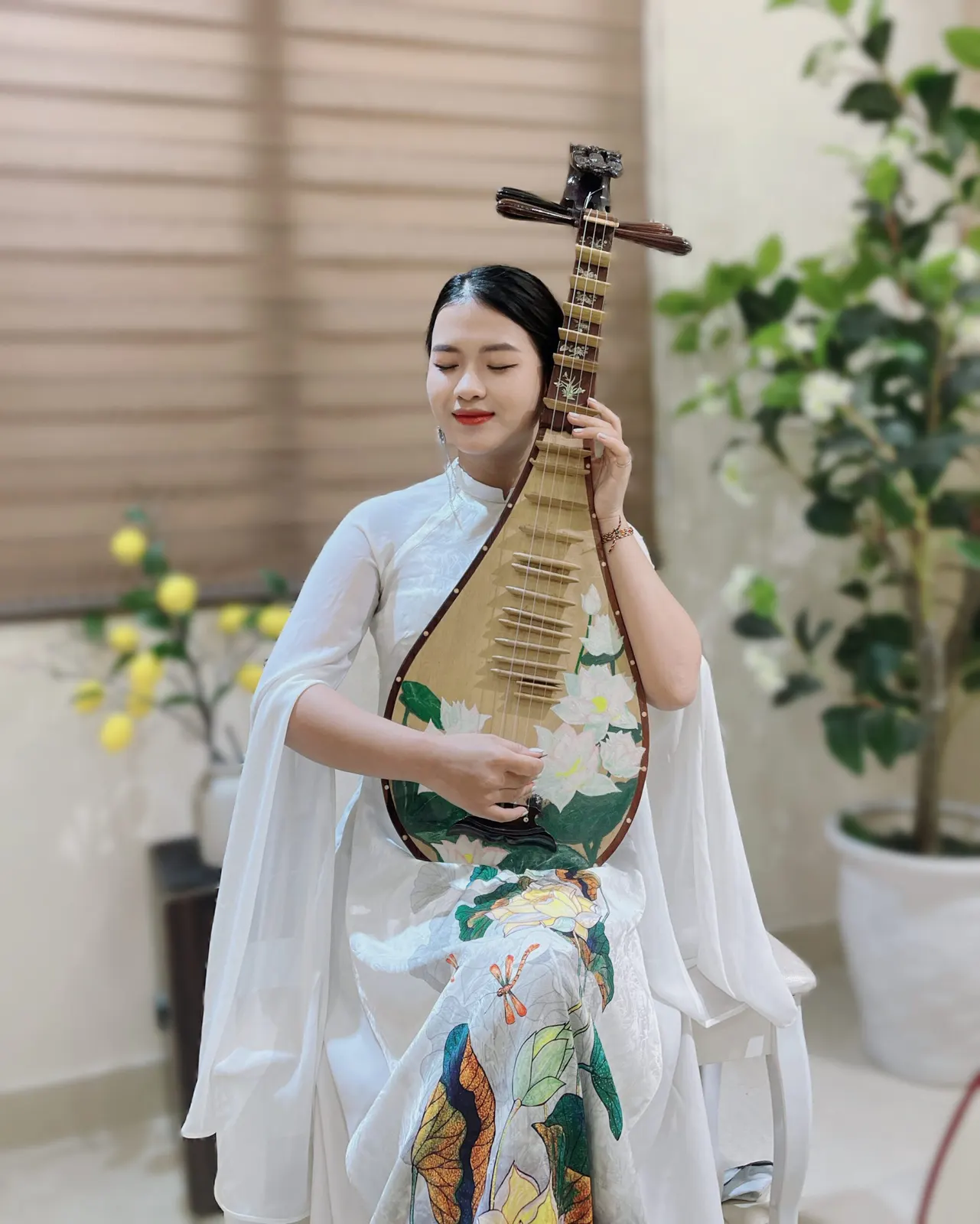 Người chơi đàn mặc áo dài trắng hoạ tiết sen, nhắm mắt ôm cây đàn tỳ bà vẽ hoa sen