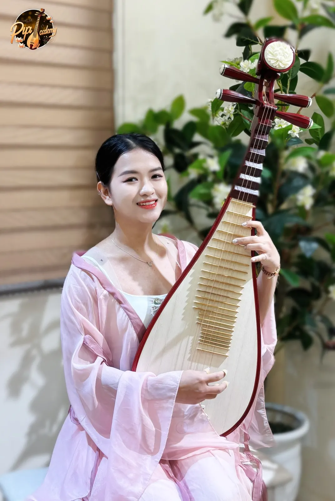 Người chơi đàn mặc áo lụa hồng mỉm cười, ôm cây đàn tỳ bà gỗ đỏ có đầu đàn chạm hoa