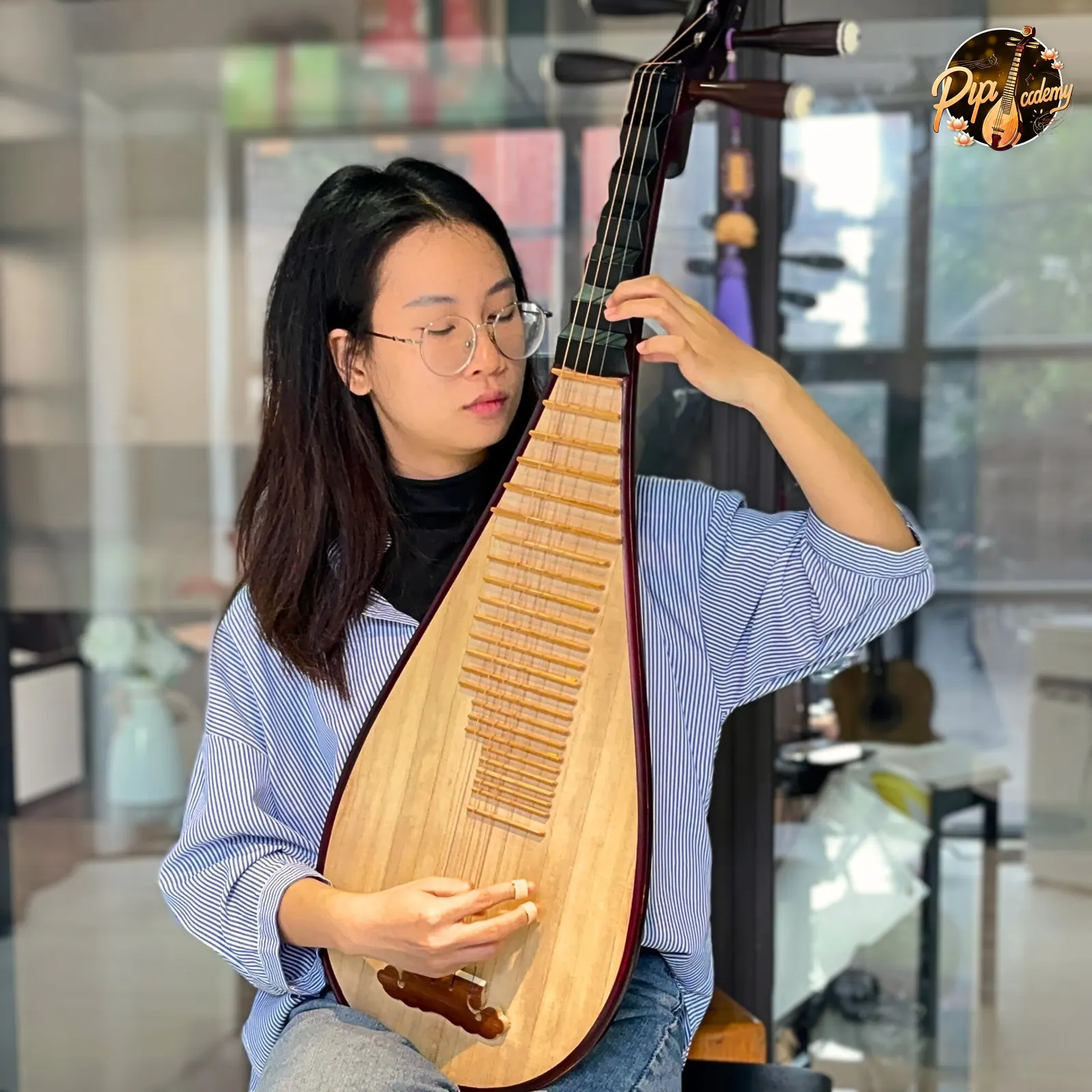 Người chơi đàn đeo kính, mặc sơ mi kẻ sọc xanh, nhắm mắt gảy đàn tỳ bà