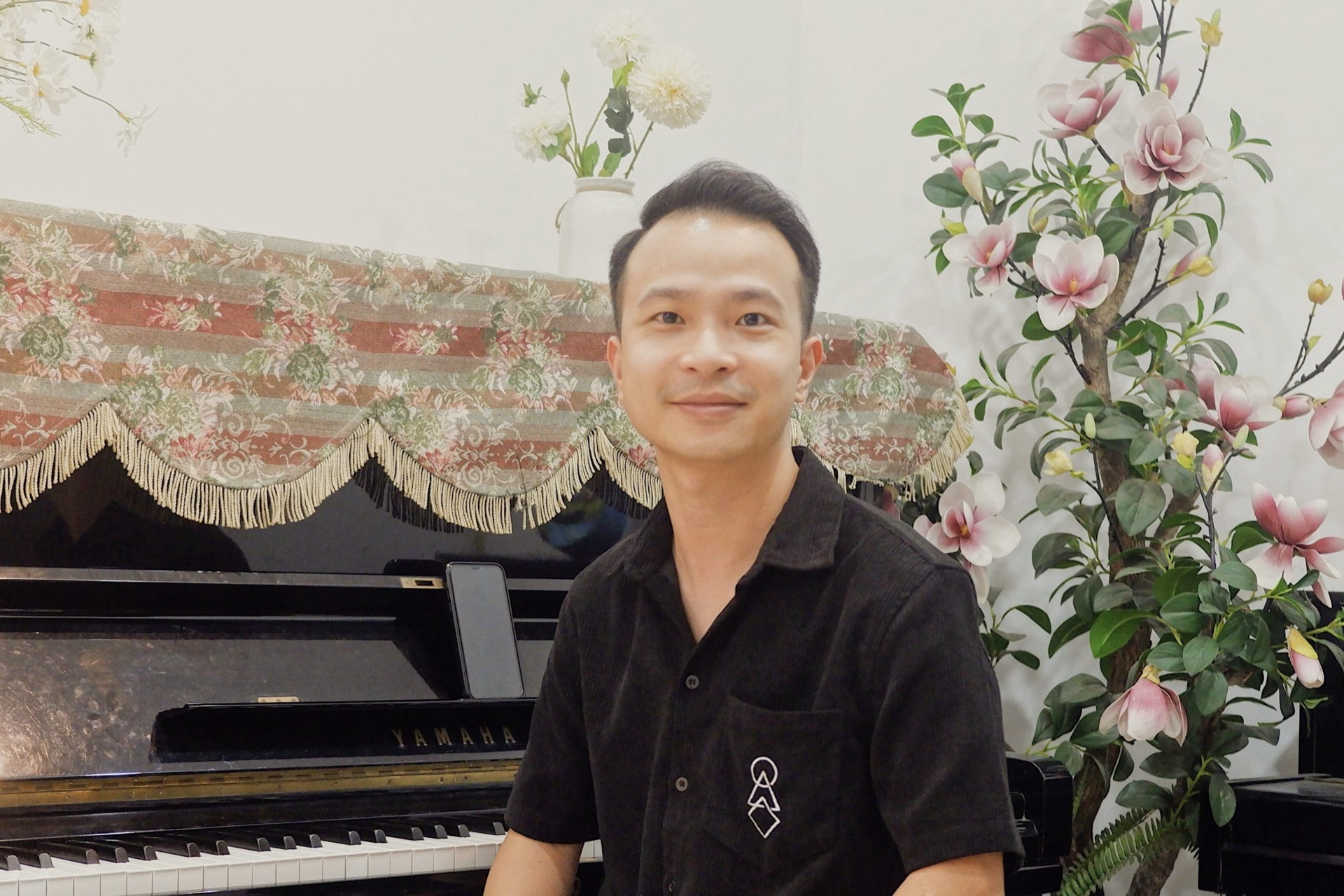 Thầy Bùi Hà Sơn, Trưởng Bộ môn Thanh nhạc Voice Academy, áo sơ mi đen, ngồi trước đàn piano Yamaha