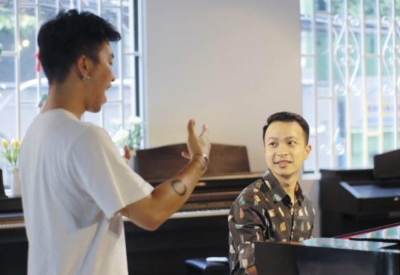 Giáo viên ngồi bên đàn piano mỉm cười nhìn học viên áo trắng đang hát
