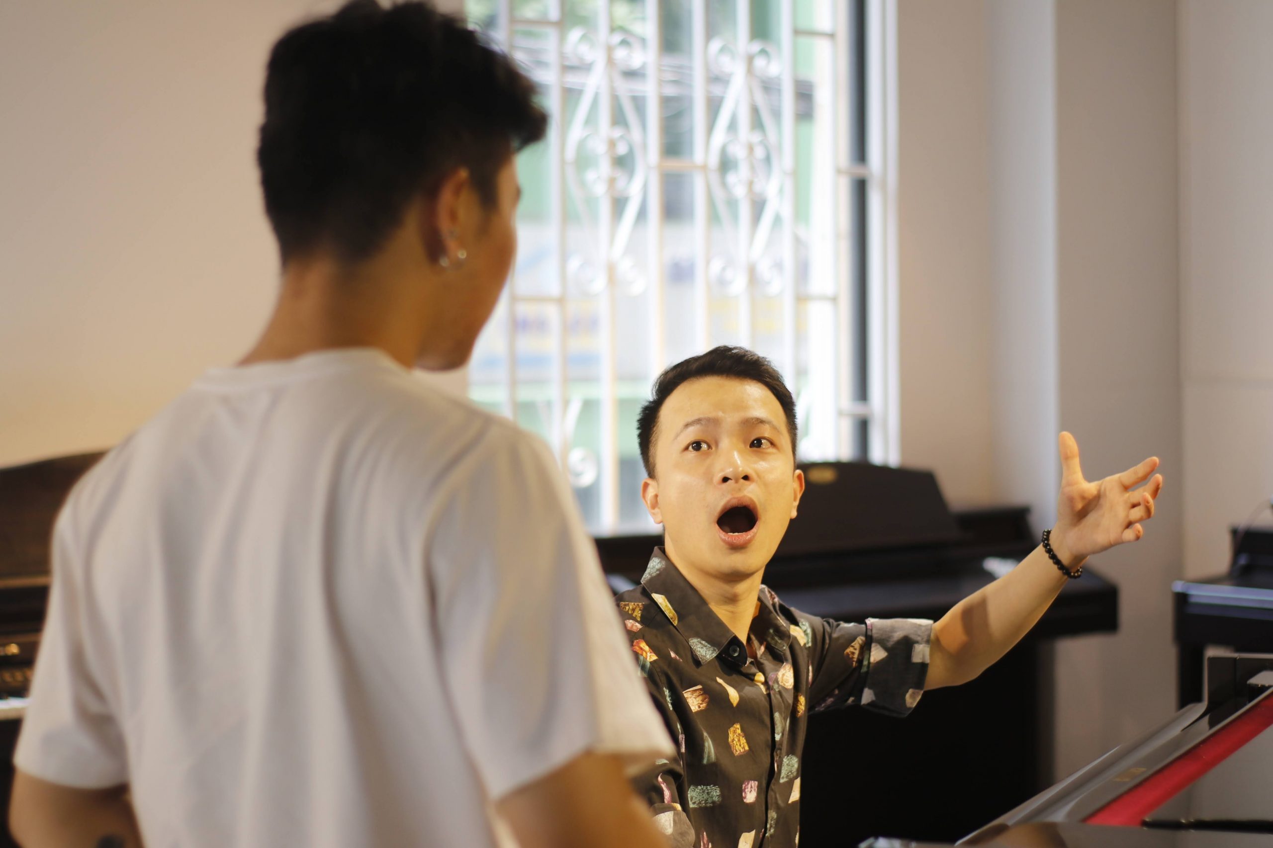 Giáo viên ngồi bên đàn piano, mở rộng khẩu hình và dang tay làm mẫu cho học viên áo trắng trong giờ luyện thanh